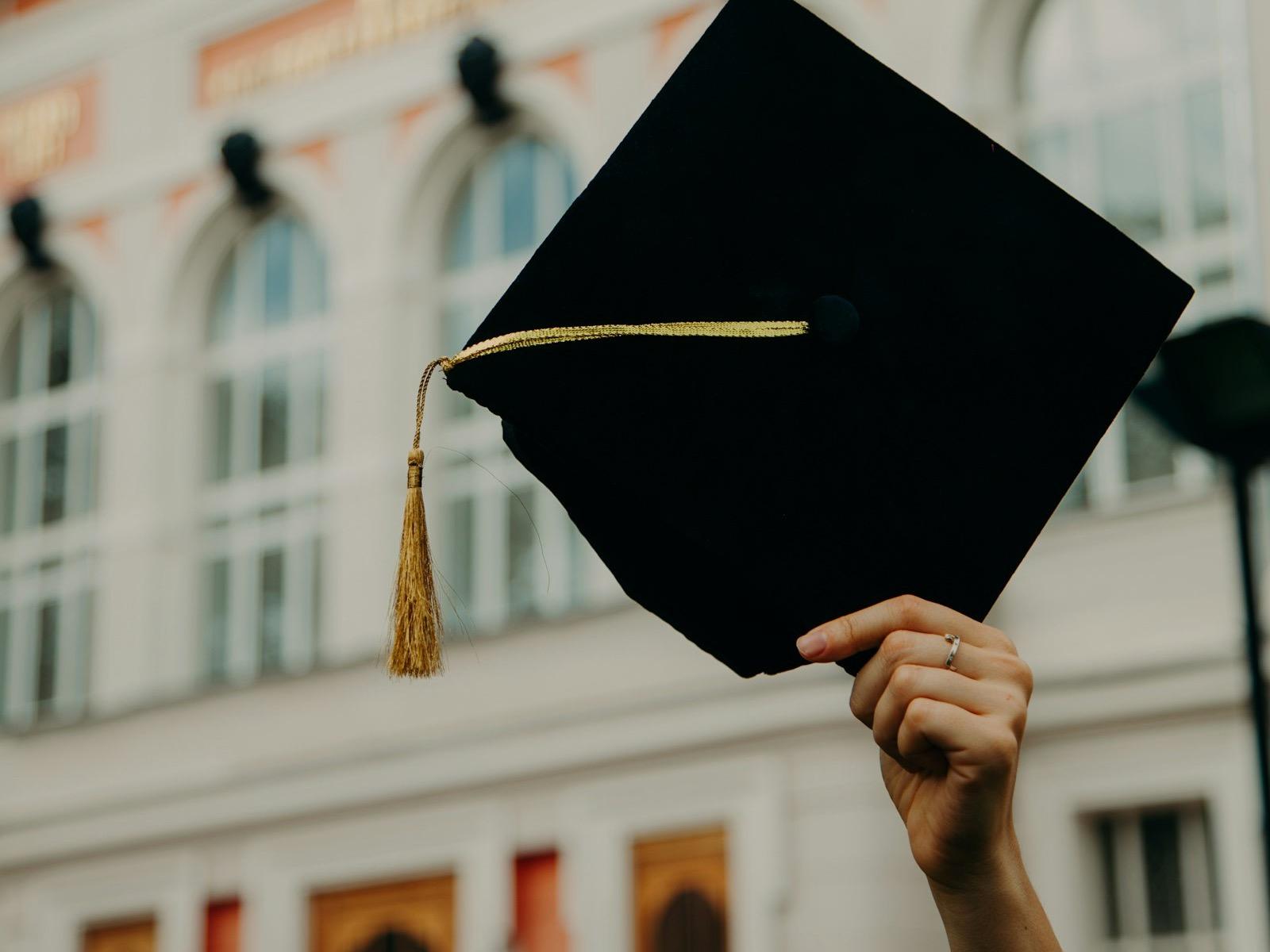 Graduate holding a black mortarboard with a gold tassel up in front of a classic building facade