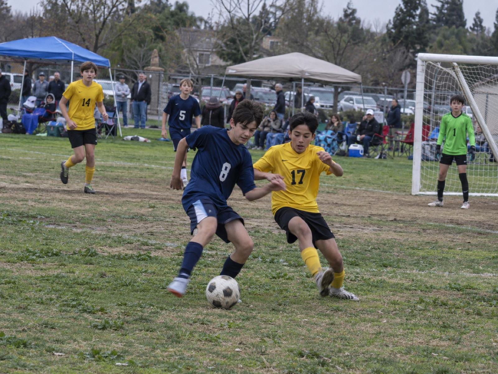 Two youth club soccer players in navy and yellow jerseys contest the ball at a Sacramento-area park as parents watch from the sideline