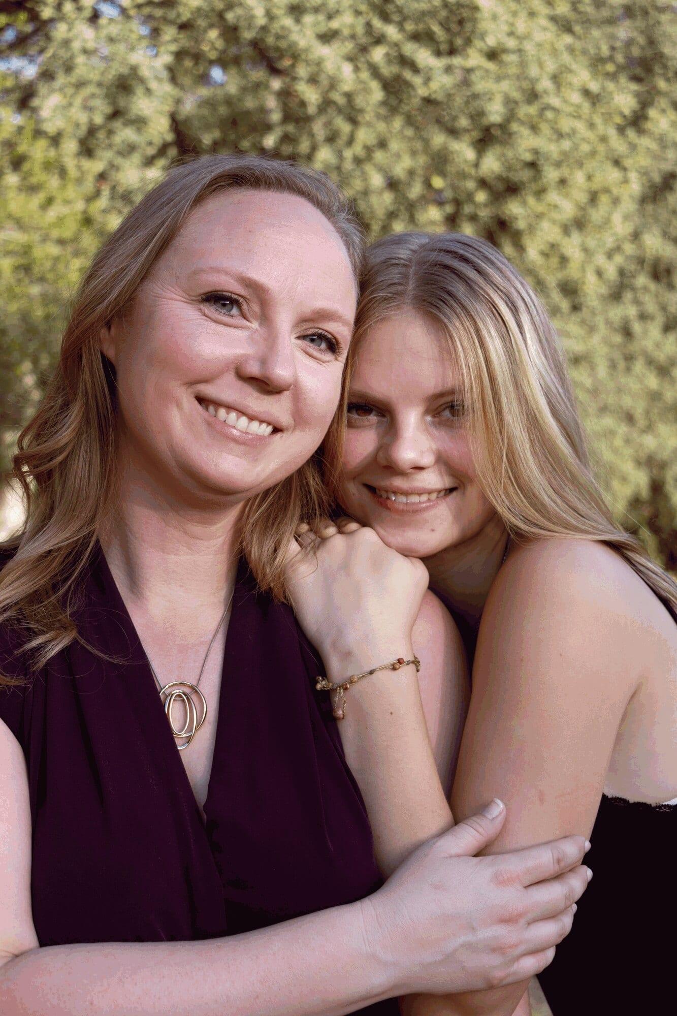 Smiling mother and teen daughter embracing cheek to cheek in front of green trees — Sacramento family photographer