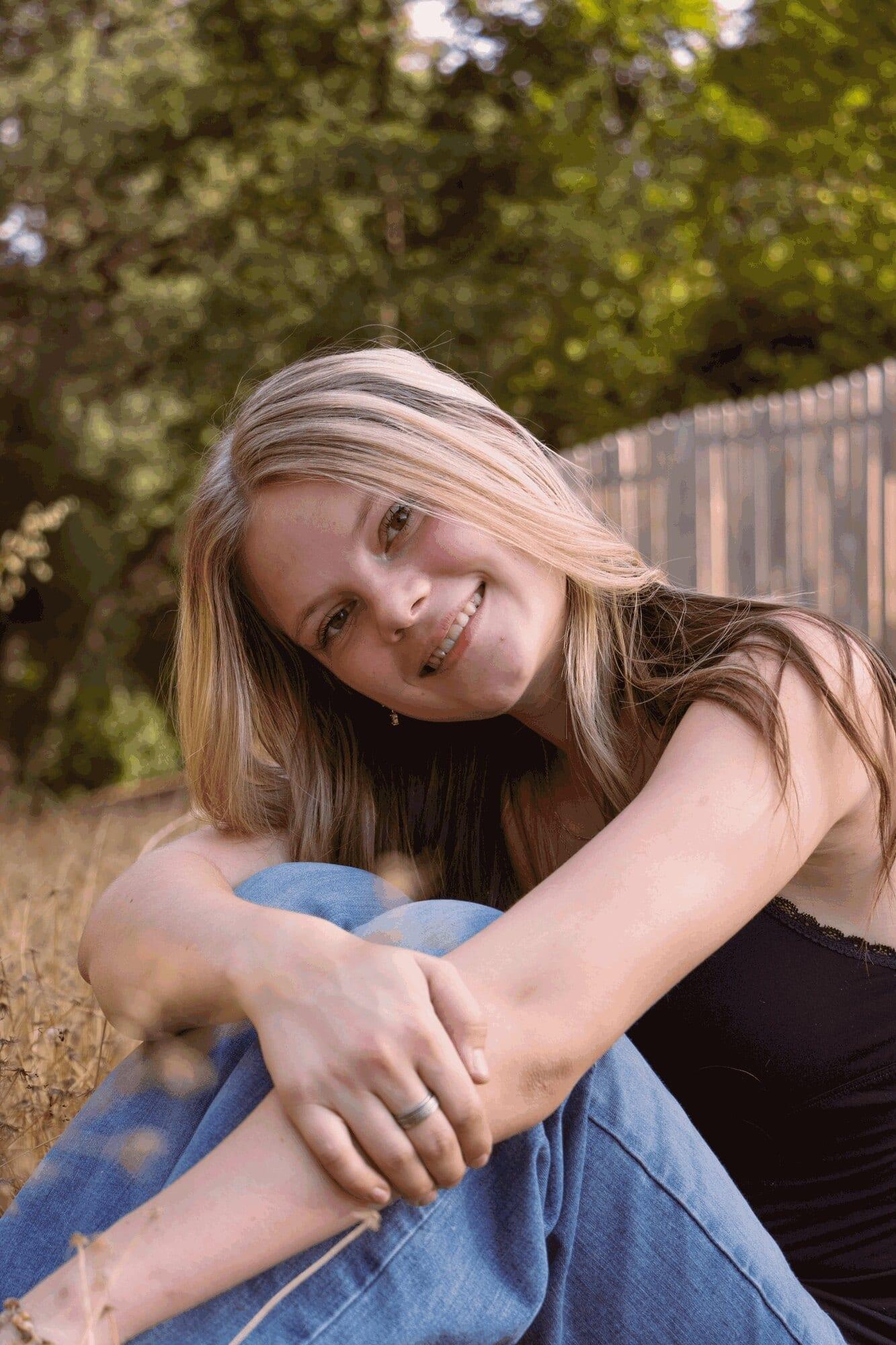 Senior girl smiling while hugging her knees in dry grass beside a wooden fence — Sacramento senior portrait photographer